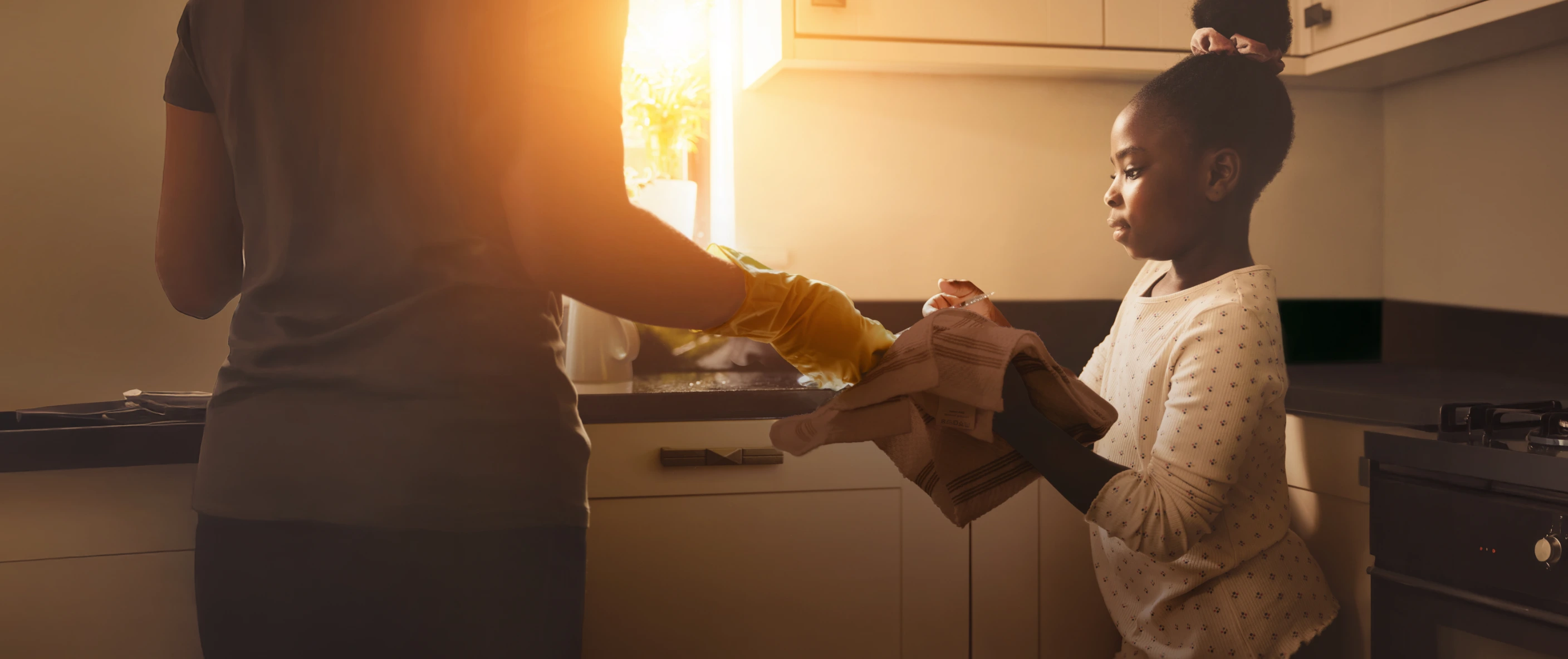 Mother handing a folded cloth to her young daughter in a warmly lit kitchen, illustrating a tender family moment